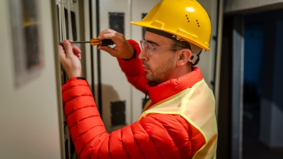 Indoors in an industrial building, a young male electrician wearing an orange down jacket, yellow safety vest, and yellow hard hat uses a screwdriver to adjust a component on an electrical panel.
