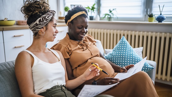 A young, mixed-race lesbian couple reviews some financial paperwork. Both women are relaxed and smiling, seated on their couch, and one of them is pregnant.