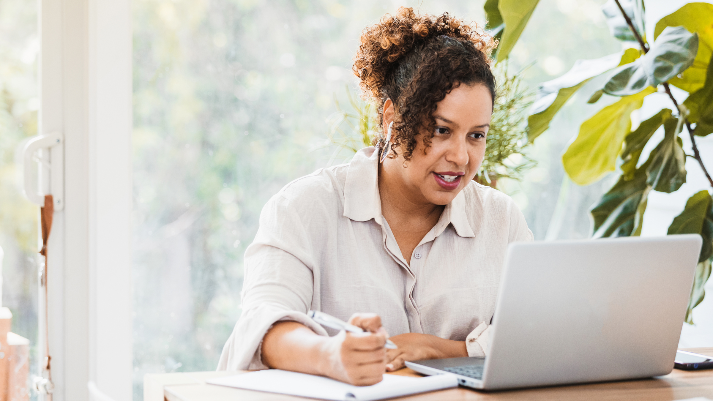 A woman attends a virtual meeting at home, via her laptop. She's seated at her dining room table, in front of a large window, and appears engaged with what she's viewing onscreen.