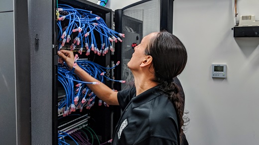 Inside a computer server room, a male Native American technician in a black shirt inspects a server panel containing dozens of blue cables.