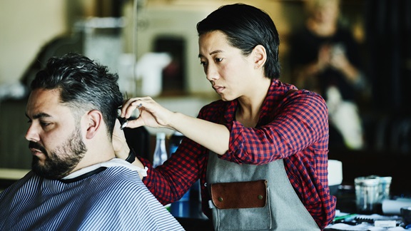 Inside a barber shop, a female barber in a red-checkered shirt and gray apron trims a male client's hair.