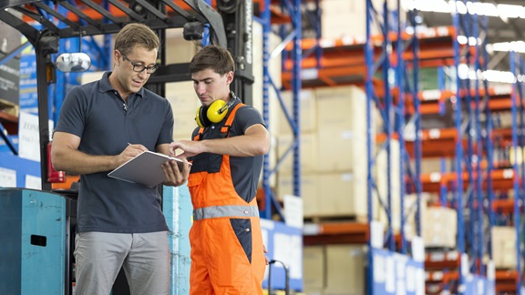 Inside a warehouse, a manager and a forklift driver are standing in the foreground and discussing some documentation that's attached to the supervisor's clipboard. The manager is a mid-adult White man wearing a navy shirt and gray pants. The forklift driver is a younger-adult White man in a navy shirt and orange overalls.