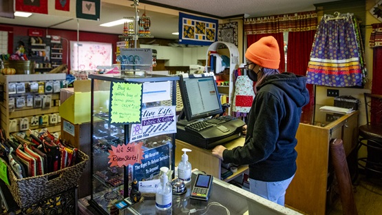 A woman in a navy sweatshirt, jeans, and orange hat works the checkout counter at Native Life, a Native-owned fabric store in Browning, Montana. The store is filled with a colorful assortment of merchandise.