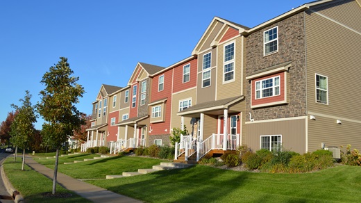 A row of recently constructed three-story townhomes with a beige-, brown-, and terra-cotta-colored exterior lines a suburban street. The sky in the background is bright blue.