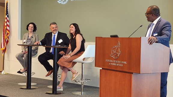 Onstage at the front of a conference room, panelists Adrienne Smith of Opportunity Finance Network, Pete Upton of the Native CDFI Network, and Lakota Vogel of Four Bands Community Fund are seated on chairs to the left of a podium, where moderator Michou Kokodoko of the Federal Reserve Bank of Minneapolis stands.