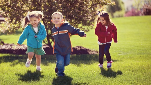In a park-like setting on a sunny day, three preschool-age children (two girls and a boy) run toward the camera, smiling and laughing
