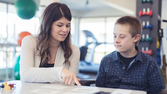 A female speech therapist works with an elementary-school-age boy in a clinic setting. They are seated at a table, looking at flash cards that are spread out before them, and the therapist is pointing out one of the cards to the boy.