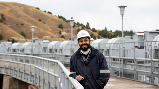 On an overcast day in what appears to be late spring or early fall, a male worker wearing a navy blue coat and a white hard hat poses and smiles on a walkway atop the Se̓liš Ksanka Ql̓ispe̓ Dam in Polson, Montana. Hilly, grassy terrain dotted with pine trees is visible behind him.
