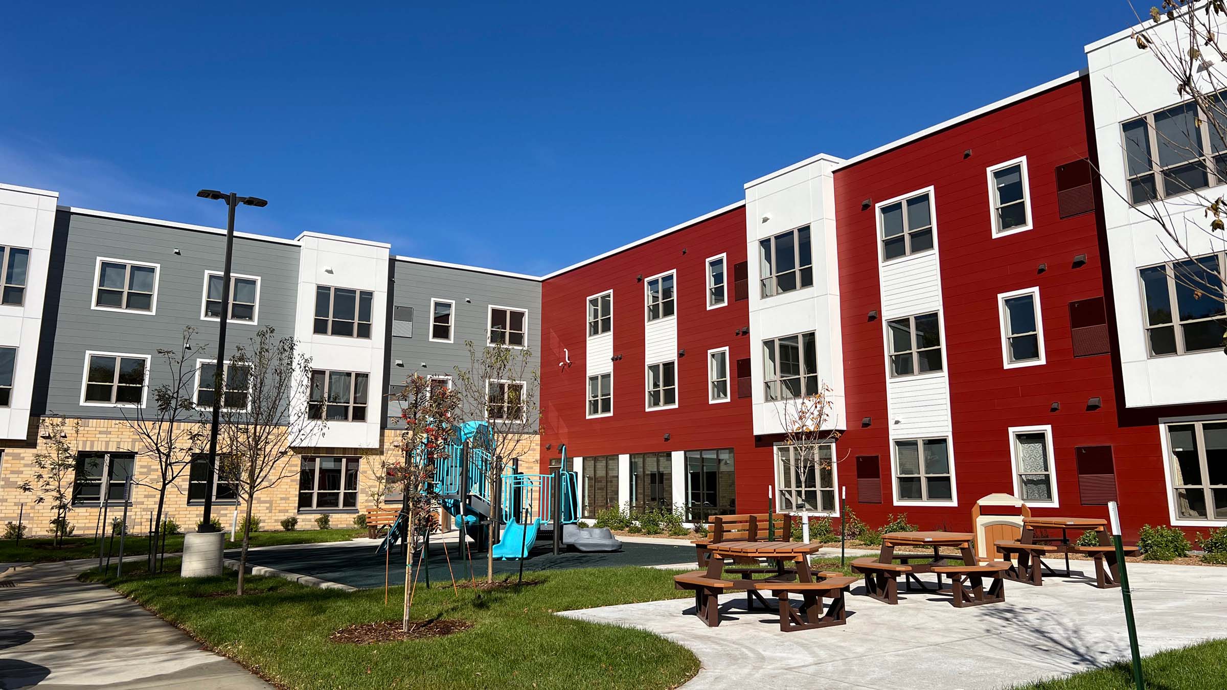An exterior view of the backside and courtyard of a newly constructed apartment complex. The building has a modern-looking facade clad in shades of red, gray, and white, with stone accents, and the courtyard features sidewalks, a cluster of light blue playground equipment, and a group of picnic tables. The day is clear and sunny.