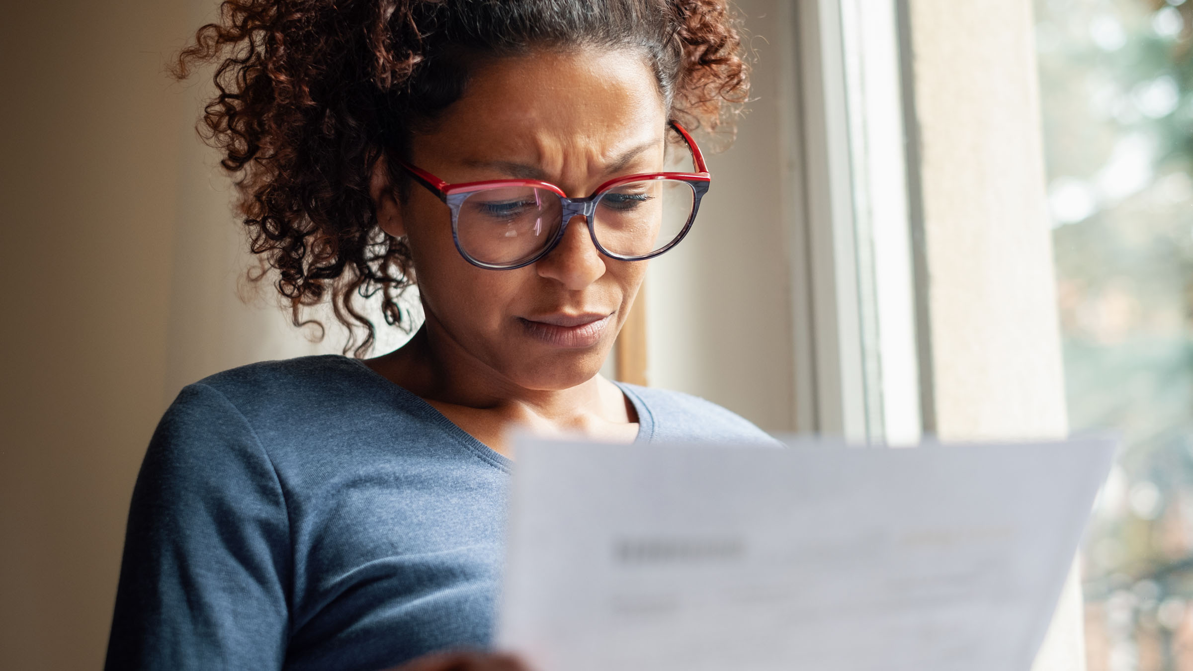 In the light of a window, a woman of color reads a form letter with a serious, concerned expression on her face.