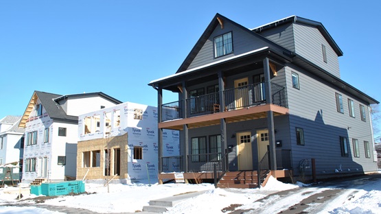 Three two-story multifamily buildings in various stages of completion on a residential street in Minneapolis. The building in the middle is completed and has gray siding, black trim and yellow doors. The building in the middle is still being framed, and the building in the background is framed and wrapped but not yet sided. It's a clear, sunny, day and the ground is snow-covered.