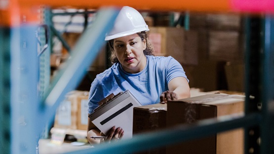 Framed by a gap in the shelving inside a warehouse, a female employee is seen inspecting boxes and checking their labels against a list she's carrying on a clipboard. She's wearing a light-blue, short-sleeved shirt and a white hard hat and has a neutral yet focused expression on her face.