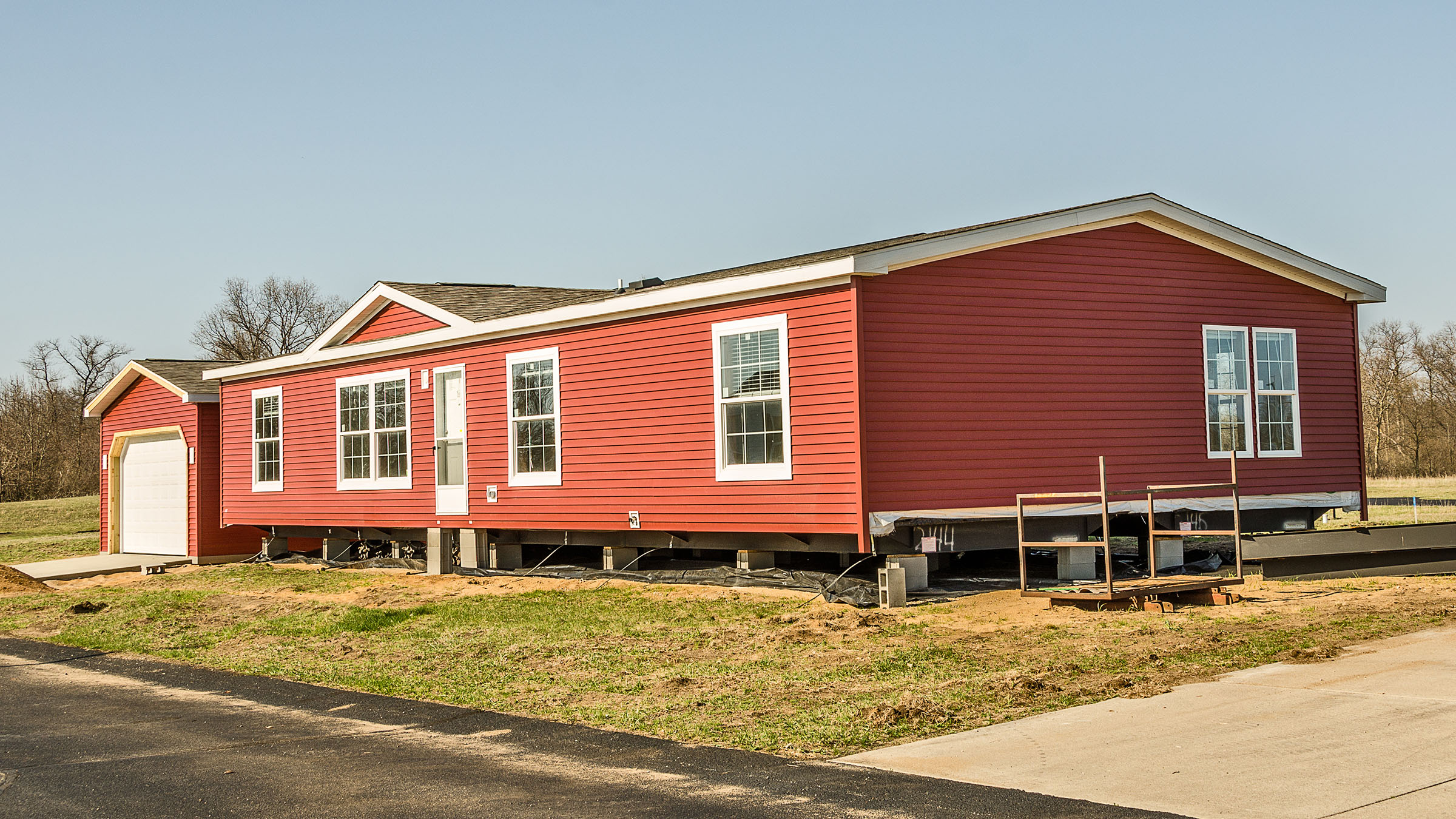 On a clear sunny day, an attractive new manufactured home with red siding and white trim sits on its lot shortly after being delivered there. The home has a garage and a newly poured driveway.