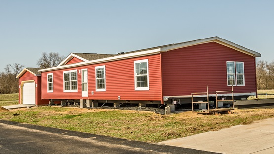 On a clear sunny day, an attractive new manufactured home with red siding and white trim sits on its lot shortly after being delivered there. The home has a garage and a newly poured driveway.
