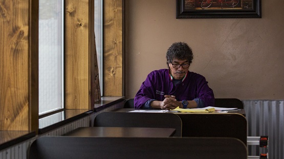 Seated in a window booth inside his restaurant, a Native American entrepreneur reviews paperwork for his business. He has a mustache and short, curly, salt-and-pepper hair, and wears glasses and a purple shirt.