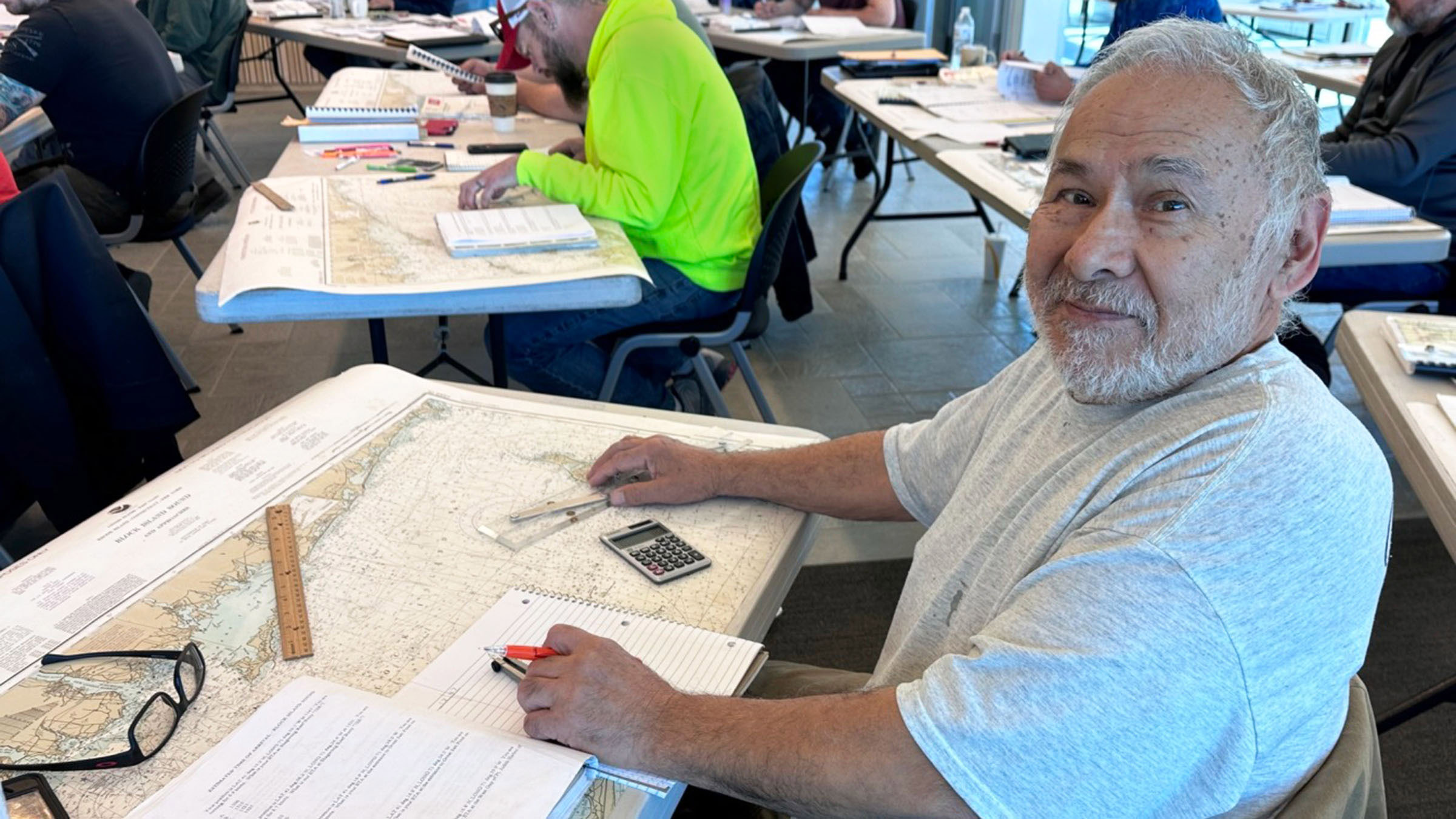 In a meeting room, a mature man in a gray tee shirt attends a charter boat captain training session. A nautical map is spread on the table in front of him, along with a workbook, calculator, navigational tools, and reading glasses. Several other class participants can be seen seated at tables in the background.