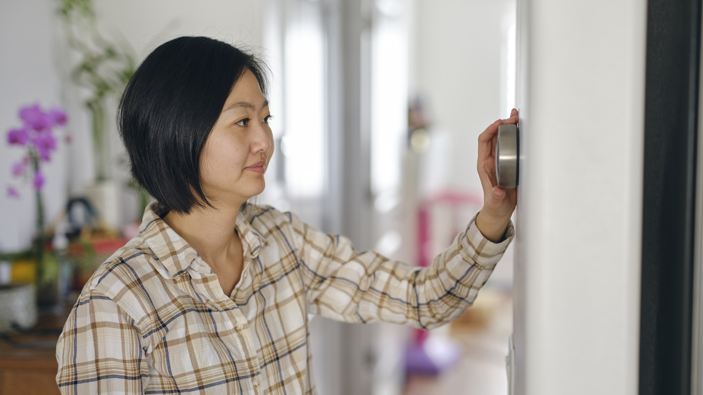 In an attractive, brightly lit home interior, a mature woman adjusts her thermostat. She is of Asian descent and is wearing a white button-down shirt with a brown and black plaid pattern.                                                                                                                                                                                                                                                                                                                                                                                                                                                                                                                                                                                                                                                                                                                                                                                                                                                                                                                                                                                                                                                                                                                                                                                                                                                                                                                                                                                                                                                                                                                                                                                                                                                                                                                                                                                                                                                                                                                                                                                                                                                                                                                                                                                                                                                                                                                                                                                                                                                                                                                                                                                                                                                                                                                                                                                                                                                                                                                                                                                                                                                                                                                                                                                                                                                                                                                                                                                                                                                                                                                                                                                                                                                                                                                                                                                                                                                                                                                                                                                                                                                                                                                                                                                                                                                                                                                                                                                                                                                                                                                                                                                                                                                                                                                                                                                                                                                                                                                                                                                                                                                                                                                                                                                                                                                                                                                                                                                                                                                                                                                                                                                                                                                                                                                                                                                                                                                                                                                                                                                                                                                                                                                                                                                                                                                                                                                                                                                                                                                                                                                                                                                                                                                                                                                                                                                                                                                                                                                                                                                                                                                                                                                                                                                                                                                                                                                                                                                                                                                                                                                                                                                                                                                                                                                                                                                                                                                                                                                                                                                                                                                                                                                                                                                                                                                                                                                                                                                                                                                                                                                                                                                                                                                                                                                                                                                                                                                                                                                                                                                                                                                                                                                                                                                                                                                                                                                                                                                                                                                                                                                                                                                                                                                                                                                                                                                                                                                                                                                                                                                                                                                                                                                                                                                                                                                                                                                                                                                                                                                                                                                                                                                                                                                                                                                                                                                                                                                                                                                                                                                                                                                                                                                                                                                                                                                                                                                                                                                                                                                                                                                                                                                                                                                                                                                                                                                                                                                                                                                                                                                                                                                                                                                                                                                                                                                                                                                                                                                                                                                                                                                                                                                                                                                                                                                                                                                                                                                                                                                                                                                                                                                                                                                                                                                                                                                                                                                                                                                                                                                                                                                                                                                                                                                                                                                                                                                                                                                                                                                                                                                                                                                                                                                                                                                                                                                                                                                                                                                                                                                                                                                                                                                                                                                                                                                                                                                                                                                                                                                                                                                                                                                                                                                                                                                                                                                                                                                                                                                                                                                                                                                                                                                                                                                                                                                                                                                                                                                                                                                                                                                                                                                                                                                                                                                                                                                                                                                                                                                                                                                                                                                                                                                                                                                                                                                                                                                                                                                                                                                                                                                                                                                                                                                                                                                                                                                                                                                                                                                                                                                                                                                                                                                                                                                                                                                                                                                                                                                                                                                                                                                                                                                                                                                                                                                                                                                                                                                                                                                                                                                                                                                                                                                                                                                                                                                                                                                                                                                                                                                                                                                                                                                                                                                                                                                                                                                                                                                                                                                                                                                                                                                                                                                                                                                                                                                                                                                                                                                                                                                                                                                                                                                                                                                                                                                                                                                                                                                                                                                                                                                                                                                                                                                                                                                                                                                                                                                                                                                                                                                                                                                                                                                                                                                                                                                                                                                                                                                                                                                                                                                                                                                                                                                                                                                                                                                                                                                                                                                                                                                                                                                                                                                                                                                                                                                                                                                                                                                                                                                                                                                                                                                                                                                                                                                                                                                                                                                                                                                                                                                                                                                                                                                                                                                                                                                                                                                                                                                                                                                                                                                                                                                                                                                                                                                                                                                                                                                                                                                                                                                                                                                                                                                                                                                                                                                                                                                                                                                                                                                                                                                                                                                                                                                                                                                                                                                                                                                                                                                                                                                                                                                                                                                                                                                                                                                                                                                                                                                                                                                                                                                                                                                                                                                                                                                                                                                                                                                                                                                                                                                                                                                                                                                                                                                                                                                                                                                                                                                                                                                                                                                                                                                                                                                                                                                                                                                                                                                                                                                                                                                                                                                                                                                                                                                                                                                                                                                                                                                                                                                                                                                                                                                                                                                                                                                                                                                                                                                                                                                                                                                                                                                                                                                                                                                                                                                                                                                                                                                                                                                                                                                                                                                                                                                                                                                                                                                                                                                                                                                                                                                                                                                                                                                                                                                                                                                                                                                                                                                                                                                                                                                                                                                                                                                                                                                                                                                                                                                                                                 n