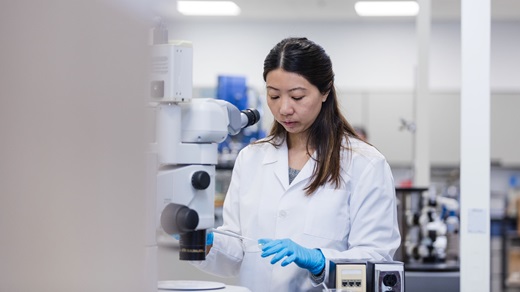 In a medical laboratory, a technician standing before a microscope uses a pipette to place a sample in a petri dish. She is Asian, has long dark hair that's partially pulled back from her face, and is wearing a white lab coat and blue latex gloves.