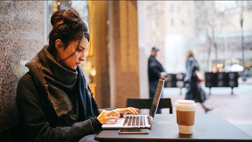 On a downtown street in a large city, a young woman works at a laptop while seated at a sidewalk table outside a coffee house. She appears in profile, wears a dark coat and scarf, and has her hair piled in a loose bun on top of her head. In the background, pedestrians and traffic are visible but out of focus.