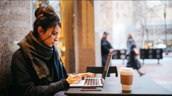 On a downtown street in a large city, a young woman works at a laptop while seated at a sidewalk table outside a coffee house. She appears in profile, wears a dark coat and scarf, and has her hair piled in a loose bun on top of her head. In the background, pedestrians and traffic are visible but out of focus.