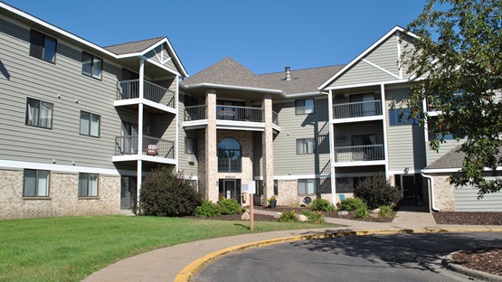 A three-story apartment building  constructed in 1989 is pictured on a clear, sunny fall day. The building has beige siding, tan brick accents, white trim, and a brown roof. 