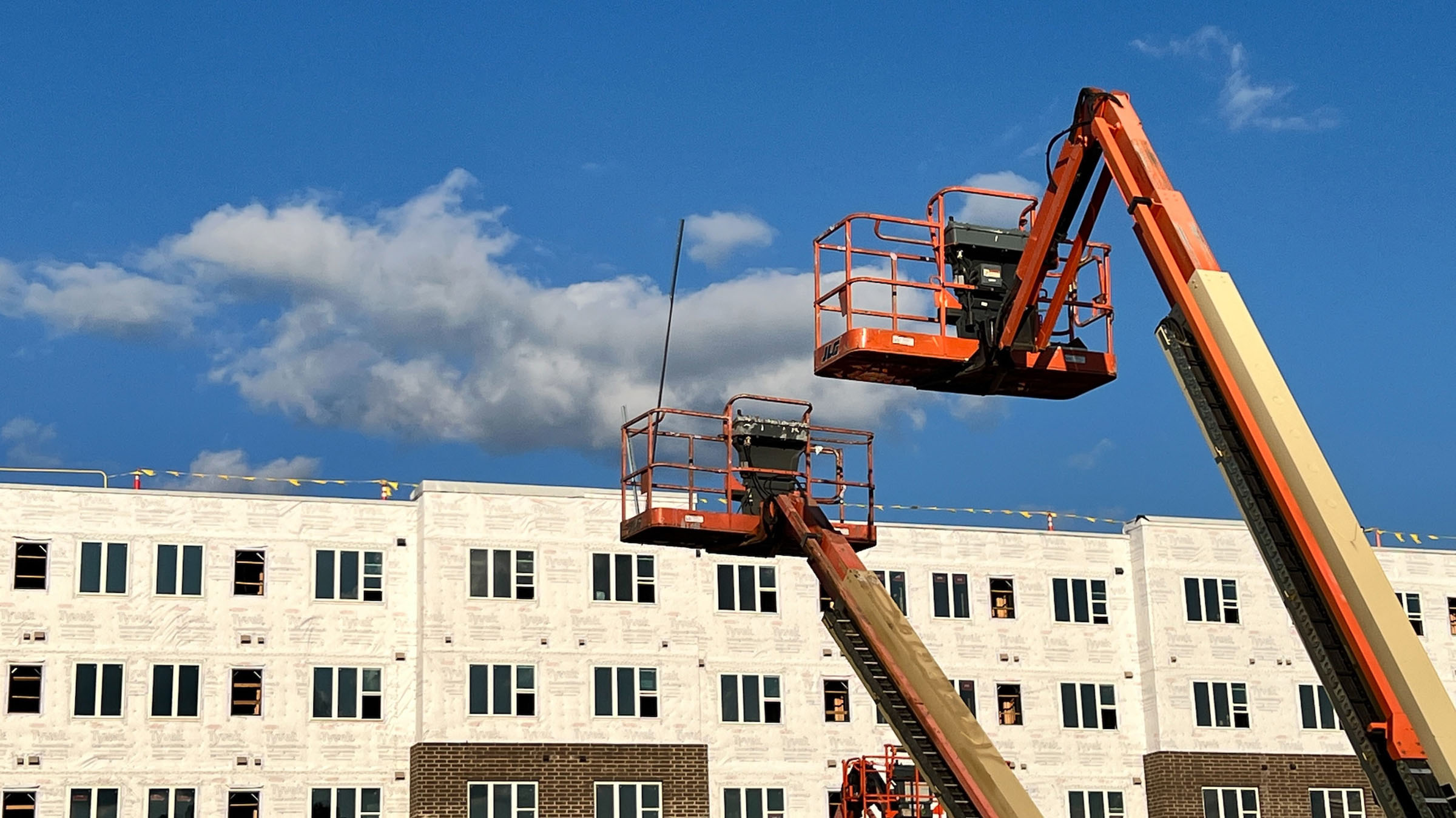 At a construction site, the baskets and arms of two orange-colored boom lifts appear in the foreground, and a partially finished apartment complex covered in white building wrap appears in the background. Blue sky with a few isolated clouds is visible above the roof of the building.