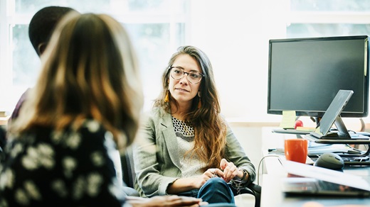 In a brightly lit office space, two clients and a lending professional sit and meet at a work station. The two clients are out of focus in the foreground, seen from behind, faces not visible, while the lending professional is in focus in the background. She has glasses and long brown hair and wears a light green blazer and blue pants. She has an attentive expression as she listens.