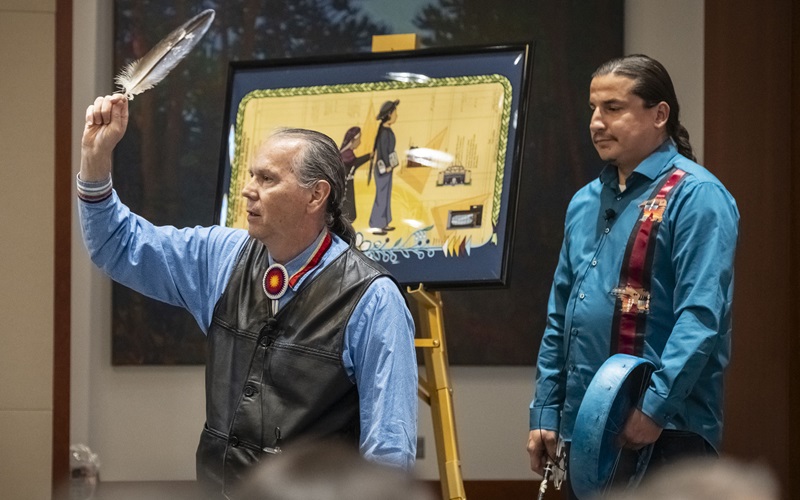At the start of the Center for Indian Country Development 10th Anniversary and Data Summit event on October 8–9, 2025, Reid Raymond and Zacheriah Red Bear provide an invocation and drum song. Raymond stands at left, speaking while holding up a large feather, and Red Bear stands behind him to the right, holding a drum.