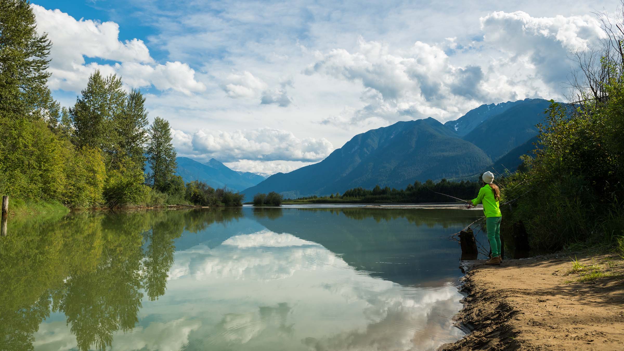 On the bank of a picturesque river in Canada, a First Nations woman casts her fishing rod into the water. She appears from behind, mid-foreground, and is wearing a fluorescent green jacket, green pants, brown boots, and a light-colored hat. Mountains, pine trees, and a partly cloudy sky are reflected on the river's surface.