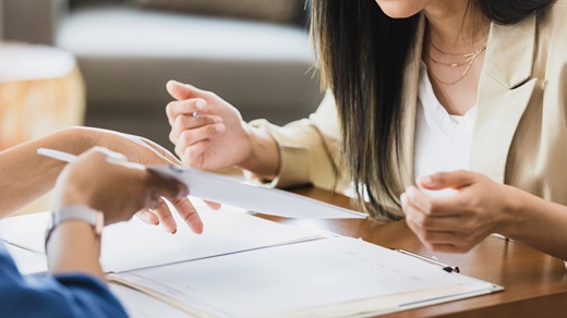 In a brightly lit office space, two women, cropped mostly out of view except for their hands, sit across from one another at a wooden desk and gesture over a folder of documents that lies open on the desktop. The woman in the foreground holds one of the documents up for the other woman to review and sign.
