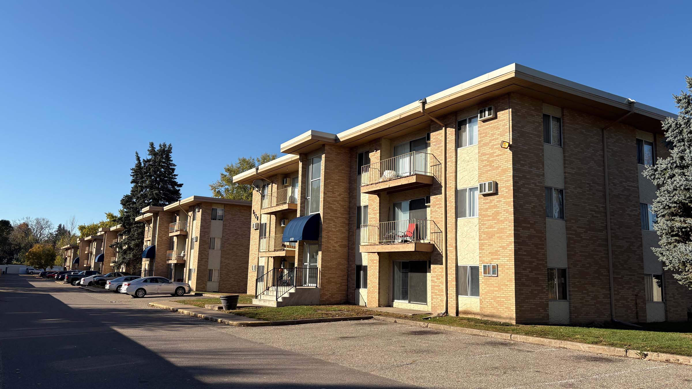 A street view of an apartment complex in Brooklyn Park, Minnesota, on a sunny, clear day. The property consists of multiple three-story, 12-unit buildings that are clad in a mixture of orange brick and cream stucco and have navy blue awnings over their front entrances. 