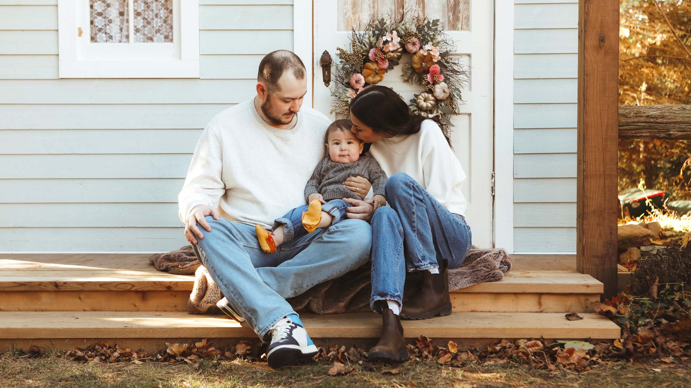 A young, straight, Indigenous couple and their baby sit on the wooden front steps of a small, light-blue house with white trim. The family members are all in jeans, with the parents wearing white sweaters and the baby wearing a gray sweater. The baby looks at the camera while the parents gaze down at the baby and the mom leans in to kiss the baby's forehead.