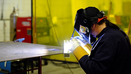 Inside an industrial building, a male worker wearing a black welding hood, navy blue long-sleeve shirt, and protective gloves bends over the edge of a metal table and welds with a small torch. The seal of the Poarch Band of Creek Indians is visible on the side of the hood. Various machines and a large, vertical, translucent yellow screen or safety shield are visible in the background.