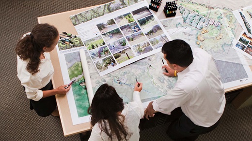 Overhead shot of three architects orlandscape designers, two women and a man, intently reviewing a set of large, colorful site plans and conceptual renderings that are spread out on a work table. One of the women is pointing out a detail in one of the plans using a pen held in her right hand.