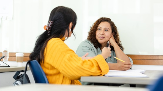 Two women engage in conversation while seated at a table in a brightly lit conference room or classroom. The first woman, in the foreground, is seen from behind. She wears a mango-colored sweater and gestures as she speaks. The woman in the background, who wears a mint-green shirt, looks at the first woman and listens attentively with her head resting on her hand.
