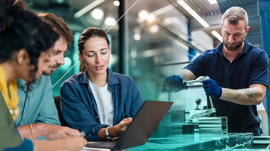 A group of college-educated workers collaborate around a computer with a green gradient behind them on the left side and a man working on a machine is on the right.