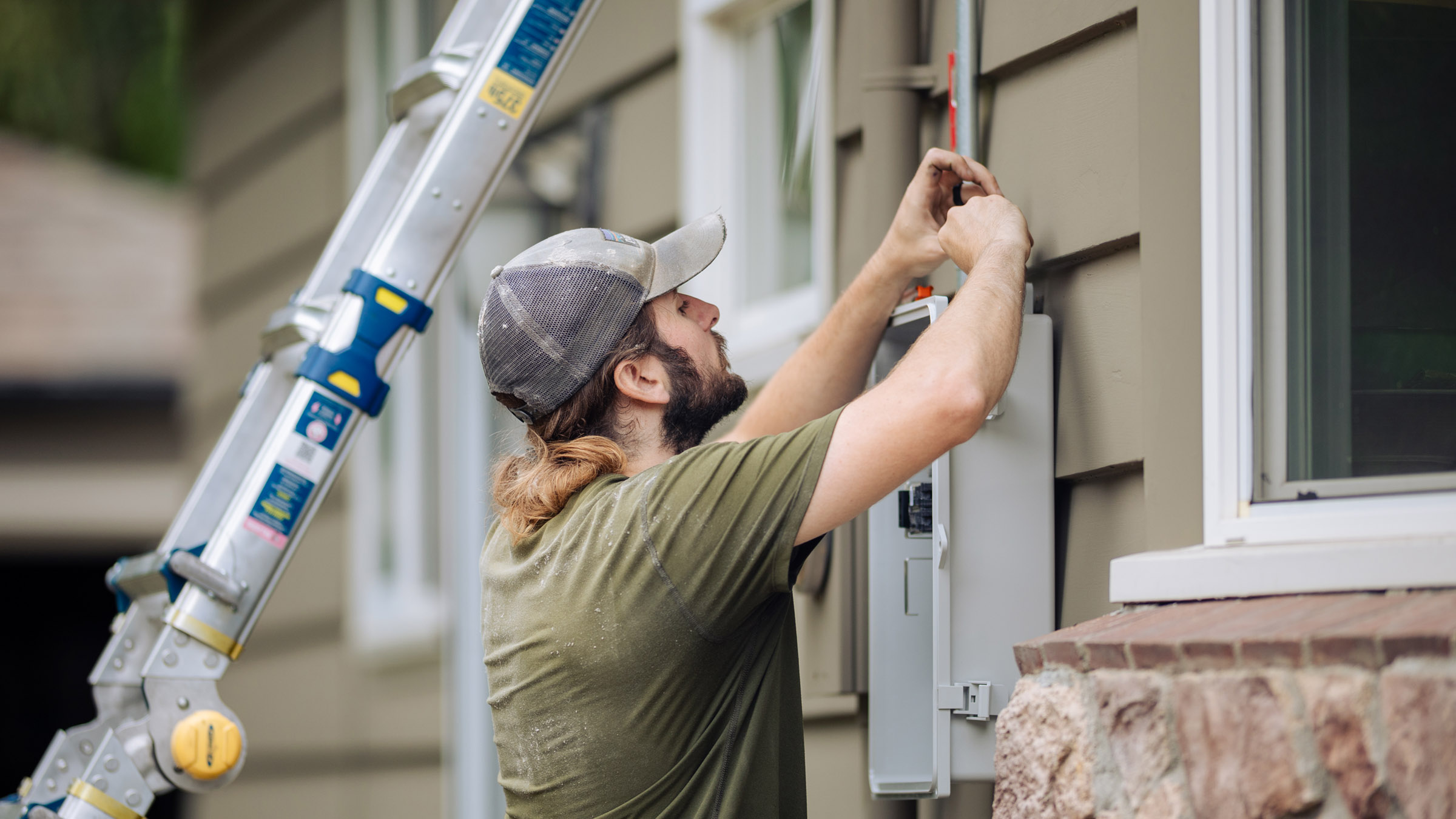 A young male electrician wearing an olive green tee shirt and gray baseball cap is seen from the side as he installs an electrical box on the exterior of a house. The house has sage green siding with white trim and stone accents. The electrician's metal ladder is visible in the background, resting against the side of the house. 