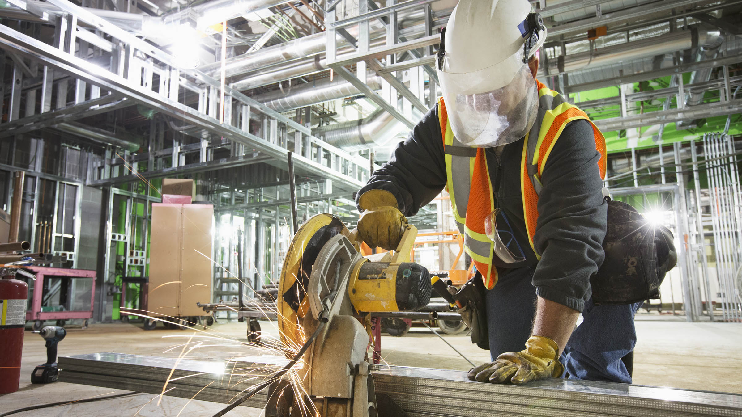 Inside the construction site of a large commercial or industrial building, with exposed steel supports and beams visible in the background, a male construction worker cuts through several lengths of steel using a power saw located in the immediate foreground. The worker is wearing a white hard hat with face shield, black sweatshirt, orange safety vest, blue jeans, and yellow work gloves. Sparks fly from the saw as it cuts the steel.