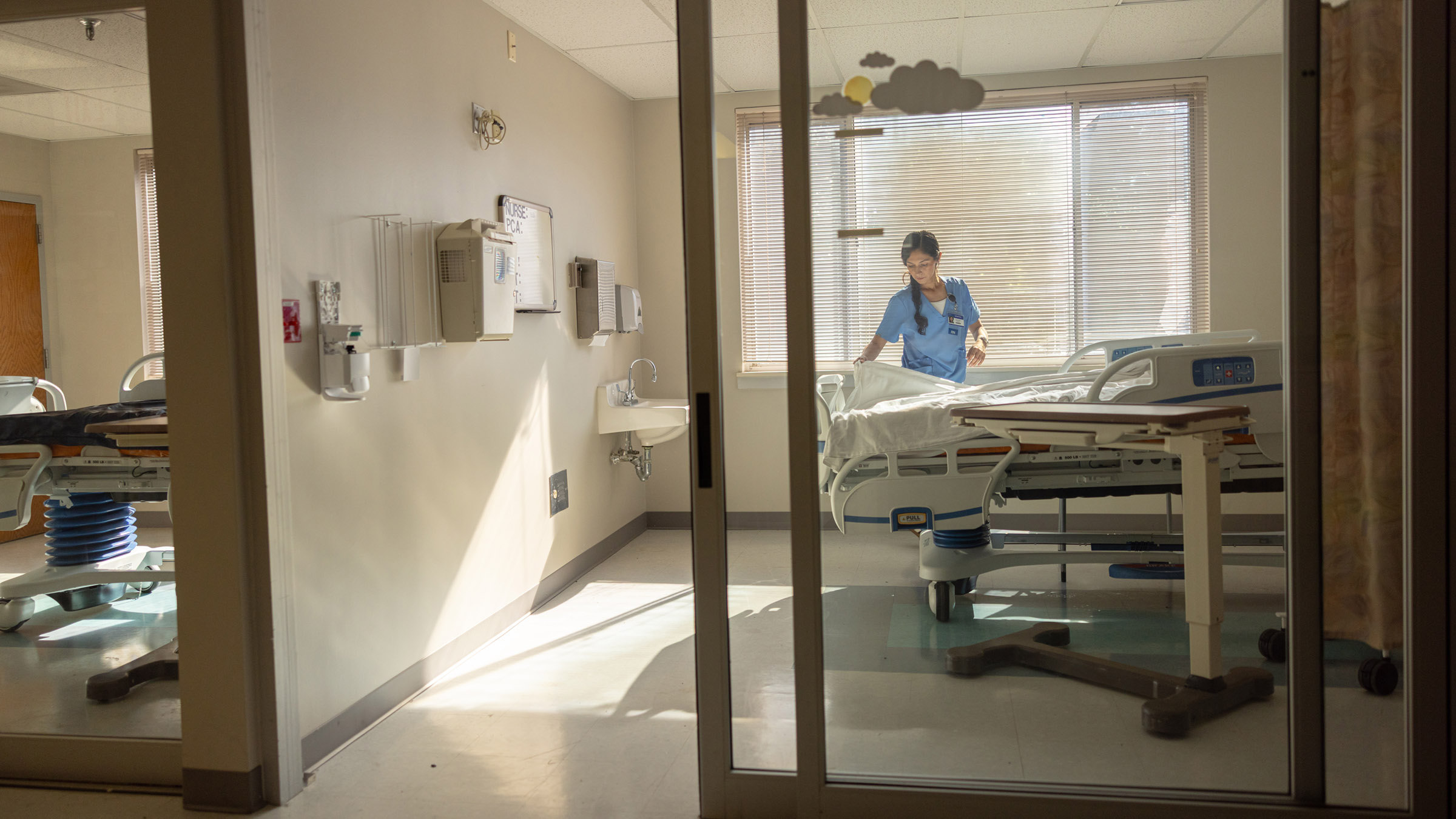 A view inside a sun-filled room in a hospital intensive care unit, where a nurse dressed in light blue scrubs adjusts the blanket as she makes the hospital bed.