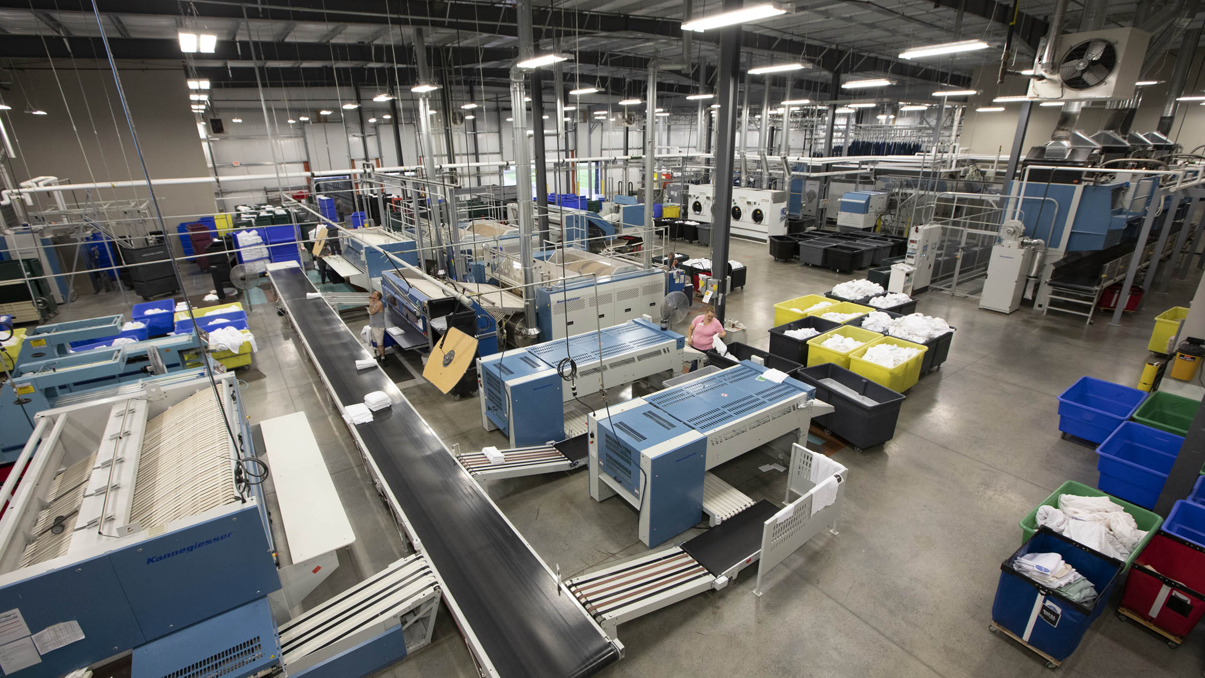 A view of the interior of Grindstone Laundry, a commercial laundry facility operated by Mille Lacs Corporate Ventures and located in Hinckley, Minnesota. The large, brightly lit space houses an array of blue industrial-sized laundry machines, bins full of linens, conveyers, and other equipment. Three workers appear in the center of the shot. The most visible of them is a woman wearing a light pink shirt.