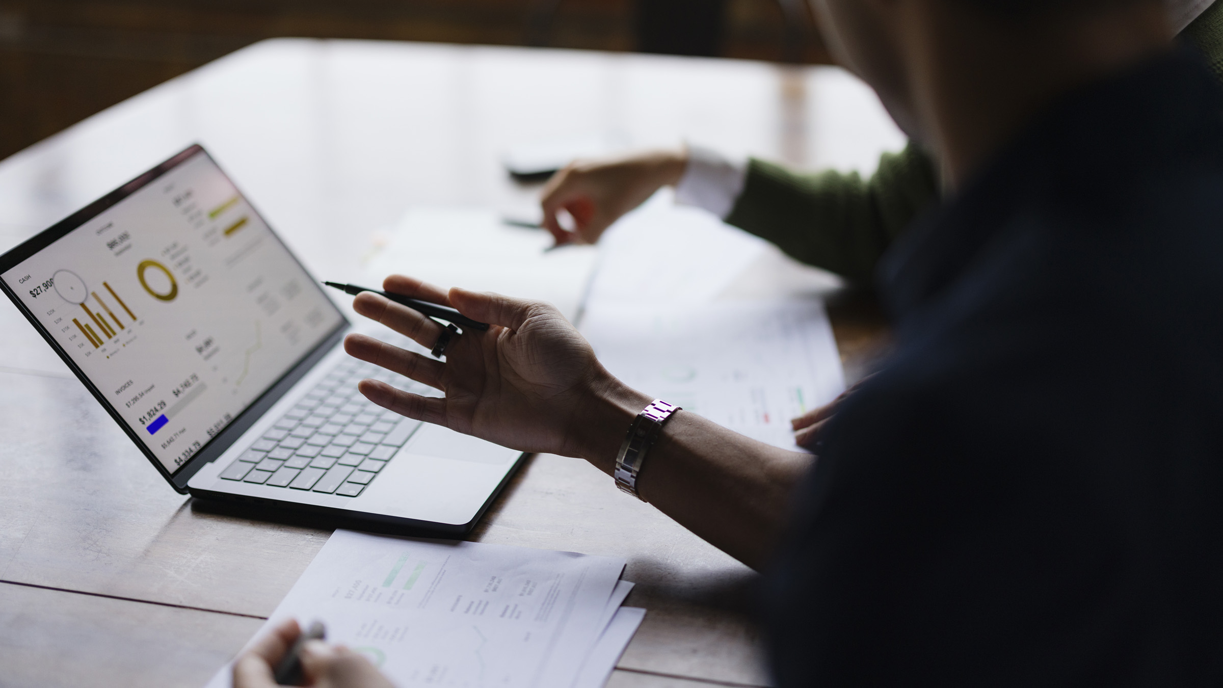 At a large wooden conference table that's lit from a nearby window, two colleagues, a man and a woman, gesture as they review financial charts and data on a laptop computer. Printouts of balance sheets lie on the table in front of them. Their heads are cut off in the shot and we only see their forearms and hands. The photo is taken in semi-profile over the shoulder of the man, who is seated in the foreground.