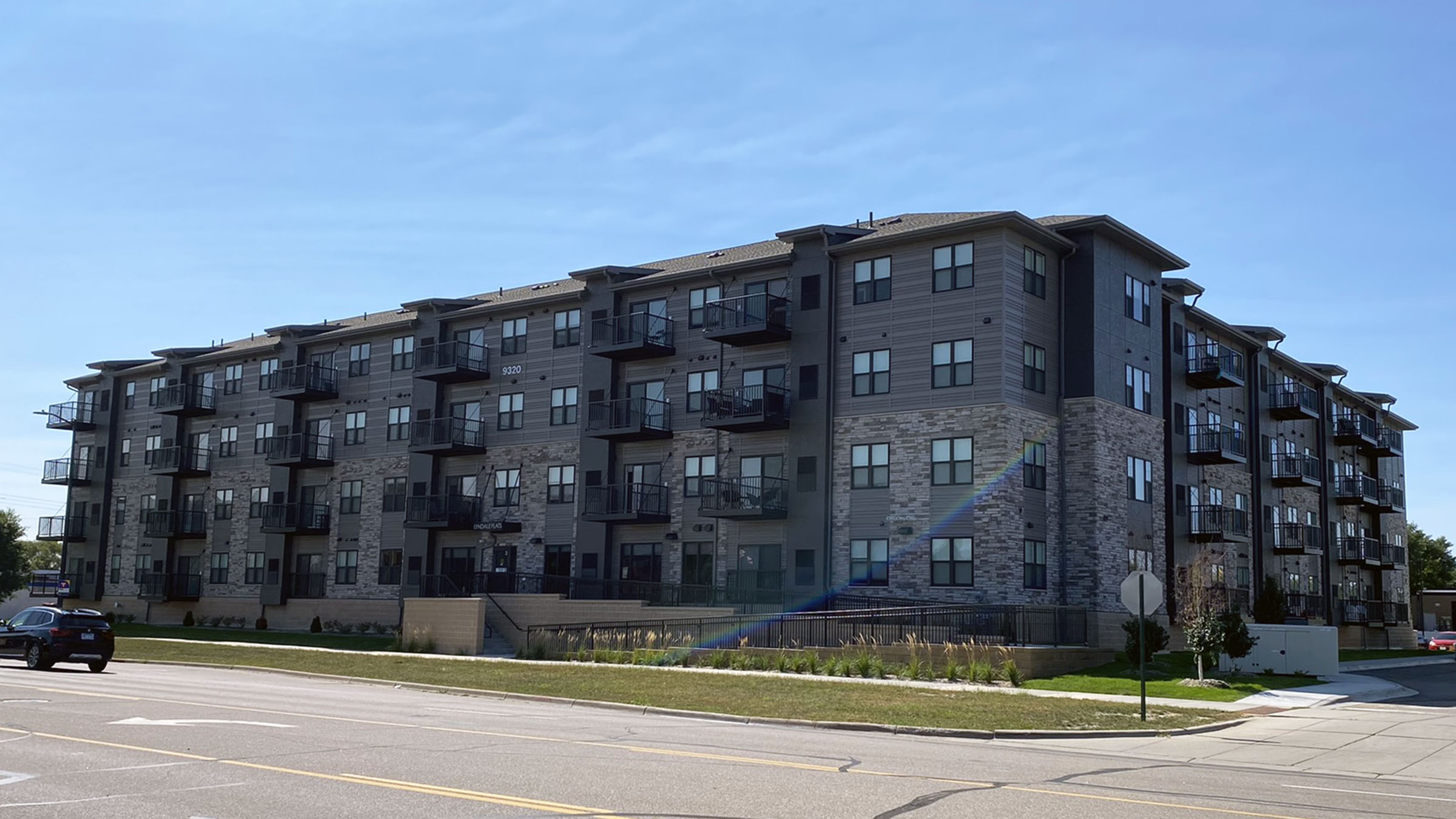 An exterior image of Lyndale Flats Apartments in Bloomington, Minnesota, shot on a clear day in spring or summer. The building is four stories high, clad in a mixture of brick and brown siding.