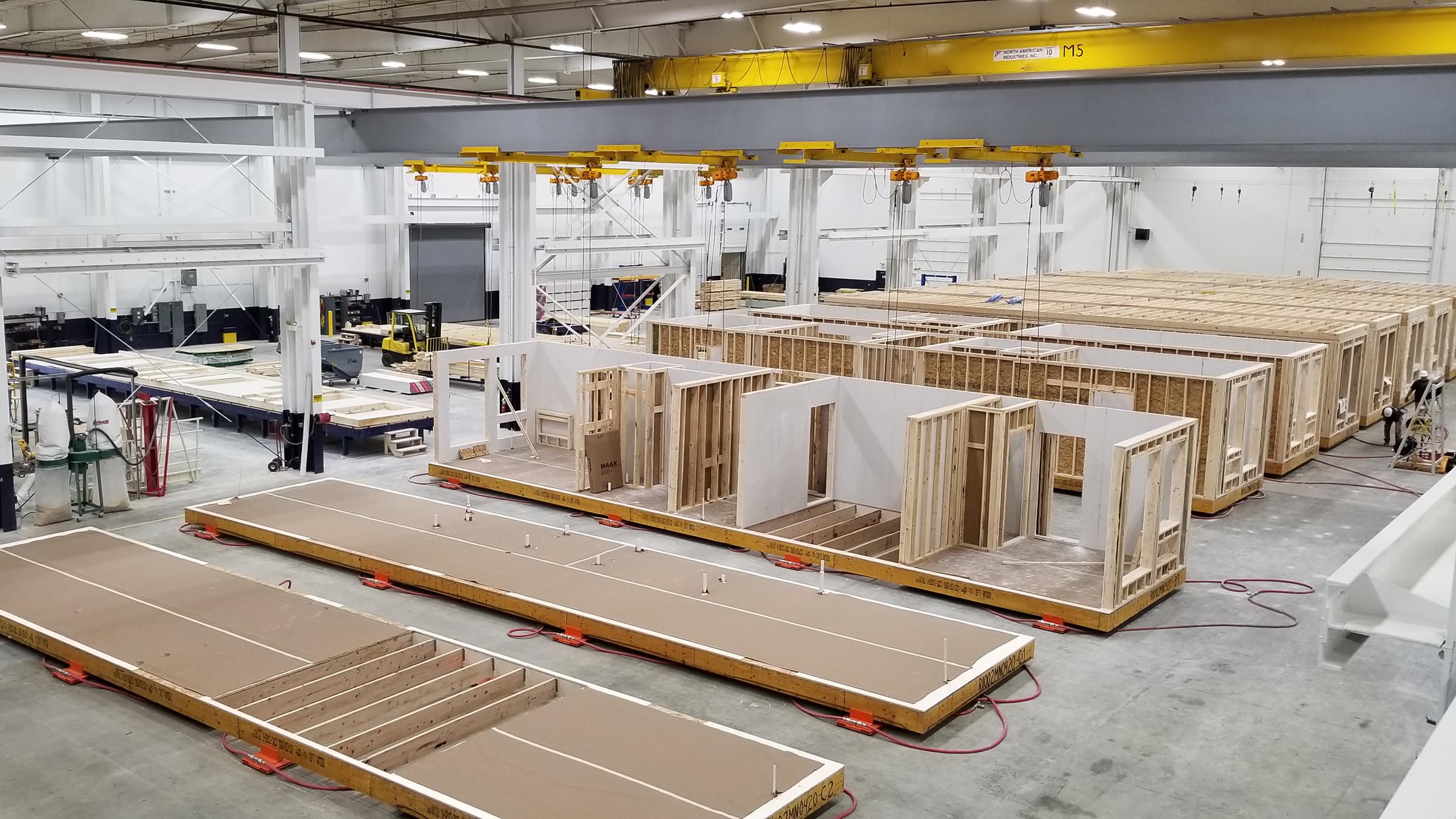 A view inside a large, brightly lit factory where units of modular housing are being constructed out of lumber and other components. The units in the foreground are only partially built and the units in the background are completely built. Two workers are visible in the upper right corner of the image..