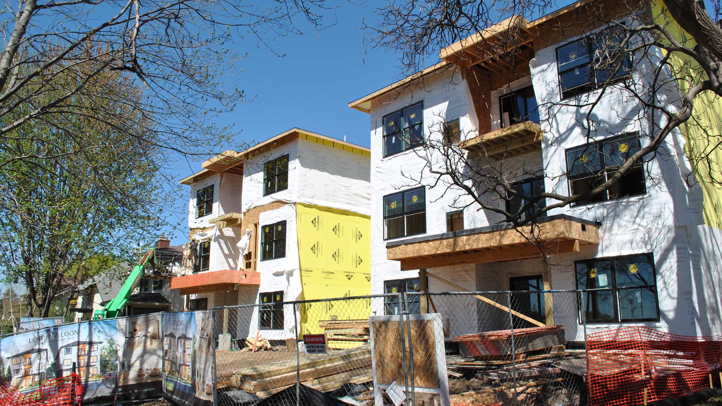 A view of two three-story, sixplex apartment buildings that are under construction on a compact urban lot in St. Paul, Minnesota, on a clear spring day. The buildings appear mostly completed except for siding and exterior finishes. They are partially wrapped in white Tyvek sheeting and the site is crowded with stacks of construction materials and temporary security fencing.