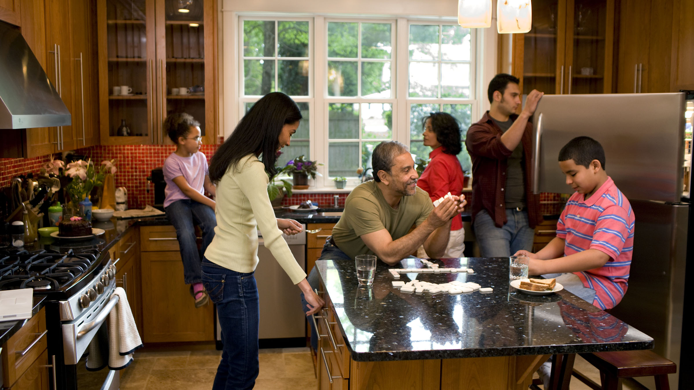 Several members of a multigenerational Latino family spend time together in their kitchen. A grandfather and grandson play dominos on the kitchen island while the mother and father begin preparing the next meal and the grandmother and young granddaughter chat by the sink, which sits below a large window. The room has a high ceiling, wooden cabinets, a red glass-tile blacksplash, black granite countertops, and stainless steel appliances.